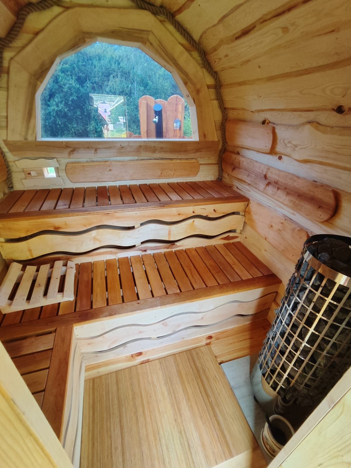 Saunaraum mit halbrundem Panoramaglas in der Rückwand, durch das man in den Garten blickt, davor zweistufige Bänke und der Harvia-Ofen